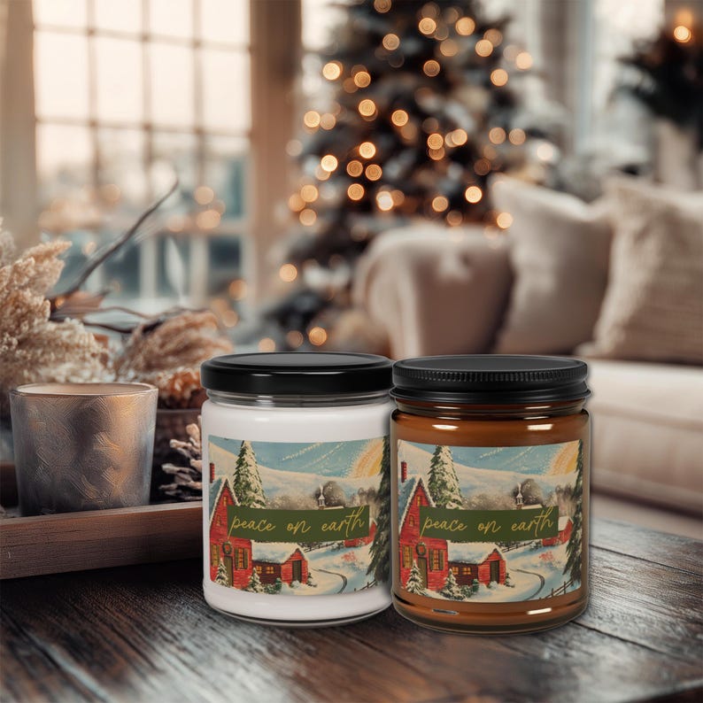 May include: Two glass jar candles with black lids. One candle is white, the other is brown. Each jar has a label with a winter scene of a red house and the text "peace on earth". The candles are on a wooden surface.