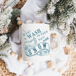 May include: White ceramic mug with a blue design that says "I want to wash my hands & hair with snow". The mug is surrounded by white and brown decorative balls and a snowy pine branch.