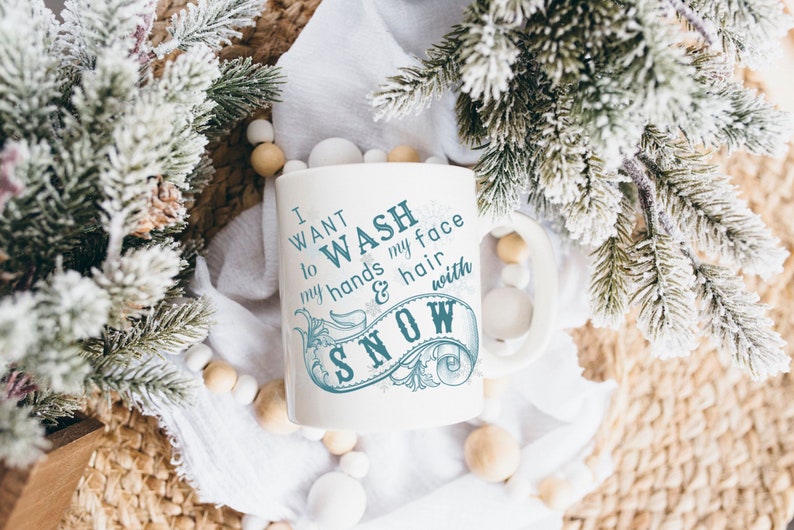 May include: White ceramic mug with a blue design that says "I want to wash my hands & hair with snow". The mug is surrounded by white and brown decorative balls and a snowy pine branch.