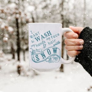 May include: White ceramic mug with a blue design that says "I want to wash my hands & hair with snow". The mug is being held by a hand wearing a black sweater in a snowy forest.