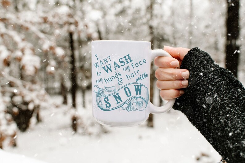 May include: White ceramic mug with a blue design that says "I want to wash my hands & hair with snow". The mug is being held by a hand wearing a black sweater in a snowy forest.