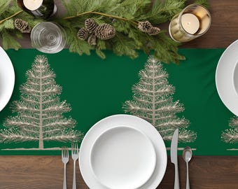 Puede incluir: Una mesa festiva con un camino de mesa verde con un diseño de árbol de Navidad plateado. Platos blancos, cubiertos y velas con piñas y vegetación completan el look navideño. La mesa está sobre una superficie de madera oscura.