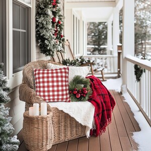 May include: A cozy porch scene with a wicker chair adorned with a red and white checkered pillow, a white pillow, and a red plaid blanket. A small wreath with red ornaments and a woven basket with candles complete the holiday decor.