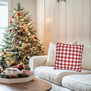 May include: A red and white checkered pillow rests on a cream-colored sofa. A Christmas tree with red ornaments and lights is in the background. The scene evokes a cozy, festive holiday atmosphere.