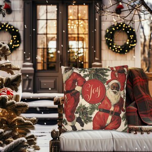 Pu&ograve; includere: Un cuscino con un design natalizio festoso che presenta due Babbo Natale in abiti rossi e una ghirlanda verde con la parola "Joy" in un cerchio. Il cuscino &egrave; su un divano bianco con una coperta a quadri rossi e neri.