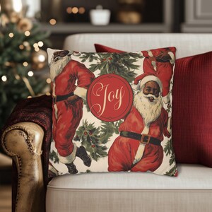 Pu&ograve; includere: Un cuscino color crema con un design natalizio festoso che presenta tre Babbo Natale neri in abiti rossi con bordi bianchi e un grande cerchio rosso con la parola "Joy" in caratteri bianchi. Lo sfondo &egrave; un motivo verde e bianco di foglie di agrifoglio e bacche.