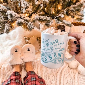 May include: White ceramic mug with a blue design that says "I want to wash my hands & hair with snow". The mug is filled with hot chocolate and marshmallows. The mug is being held by a hand with black nail polish. The mug is sitting on a white fuzzy blanket.