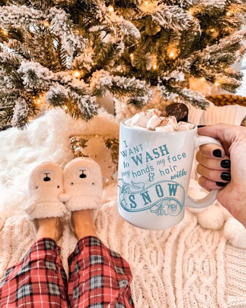 May include: White ceramic mug with a blue design that says "I want to wash my hands & hair with snow". The mug is filled with hot chocolate and marshmallows. The mug is being held by a hand with black nail polish. The mug is sitting on a white fuzzy blanket.