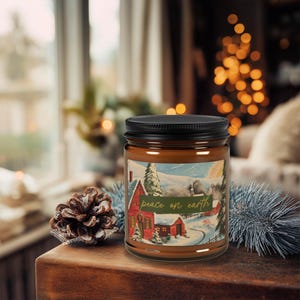 May include: A brown glass jar candle with a black lid. The label features a winter scene with a red house and the text "peace on earth" in green. A pine cone and decorative greenery are in the foreground.
