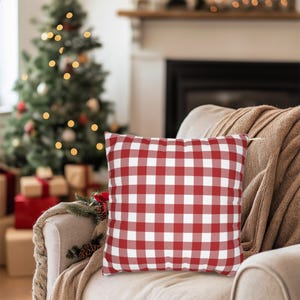 May include: A red and white checkered square pillow rests on a beige armchair, with a brown blanket draped over the arm. A Christmas tree with lights and wrapped gifts are in the background, creating a cozy holiday scene.