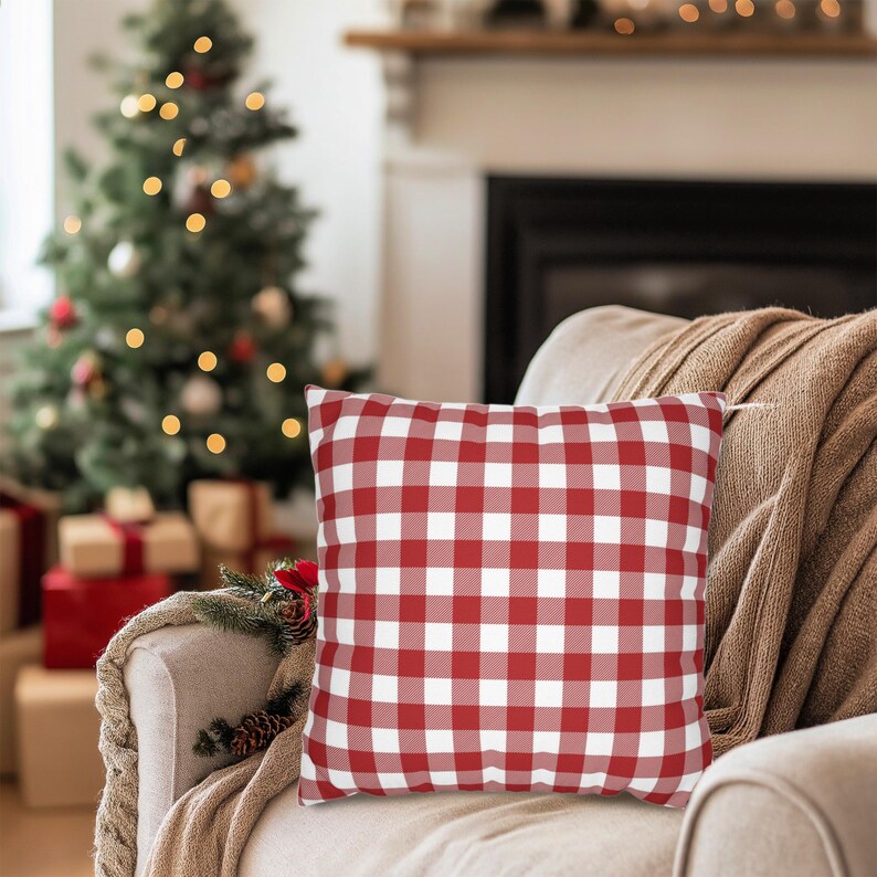 May include: A red and white checkered square pillow rests on a beige armchair, with a brown blanket draped over the arm. A Christmas tree with lights and wrapped gifts are in the background, creating a cozy holiday scene.