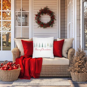 May include: A white throw pillow with a red "Merry Christmas" inscription and a row of green and blue watercolor trees. The pillow is on a wicker couch with red throw blankets and a red Christmas wreath on the wall behind it.