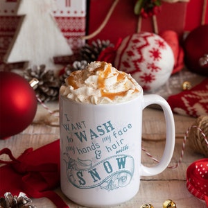 May include: White ceramic mug with a blue and white design that says "I want to wash my face and hair with snow". The mug is filled with a creamy beverage topped with whipped cream and caramel sauce. The mug is surrounded by Christmas decorations, including a red ornament, a white knitted ornament, and a pine cone.