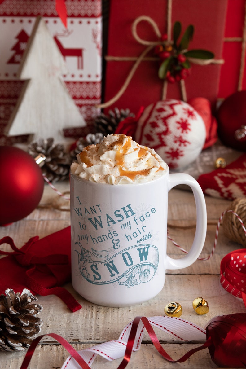 May include: White ceramic mug with a blue and white design that says "I want to wash my face and hair with snow". The mug is filled with a creamy beverage topped with whipped cream and caramel sauce. The mug is surrounded by Christmas decorations, including a red ornament, a white knitted ornament, and a pine cone.