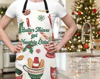Puede incluir: Delantal blanco con temática navideña con ilustraciones rojas y verdes de bastones de caramelo, galletas y tazas. El texto "Batter Thieves get Naughty Listed!" está impreso en el delantal.