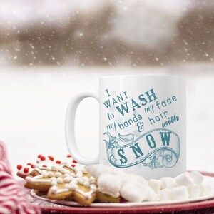 May include: White ceramic mug with a blue design that says "I want to wash my hands & hair with snow". The mug is sitting on a plate with marshmallows and gingerbread cookies.