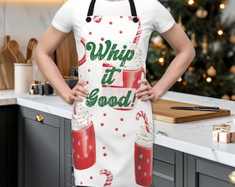 Puede incluir: Delantal de cocina blanco con un diseño navideño rojo y verde. El delantal presenta un patrón repetido de tazas con crema batida y bastones de caramelo. El texto "Whip it Good!" está impreso en verde en el delantal.