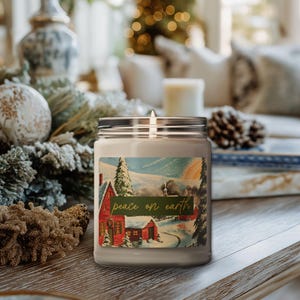 May include: A lit white candle in a glass jar with a silver lid. The label features a winter scene with a red house and the text "peace on earth". The candle is on a wooden surface, surrounded by Christmas decorations.