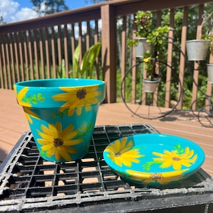 May include: A turquoise ceramic planter with a painted sunflower design. The planter has a matching saucer.