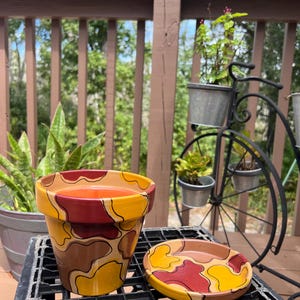 May include: A hand-painted ceramic flower pot and matching saucer. The pot and saucer feature a camouflage pattern in shades of yellow, brown, and burgundy. The pot is filled with an orange substance. The set is placed on a black metal surface.
