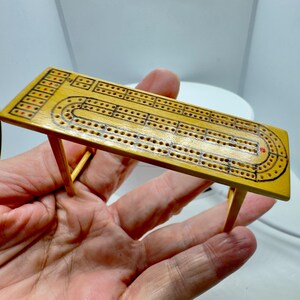 Op de afbeelding: Een klein cribbage-bord van lichtgekleurd hout met een gladde, gepolijste afwerking. Het bord heeft een rechthoekige vorm met vier dunne pootjes en is gemarkeerd met zwarte inktlijnen en kleine gaatjes voor de puntentelling, inclusief rode accenten.