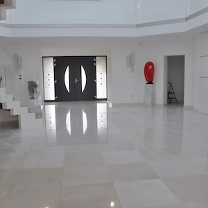 Op de afbeelding: Een modern huisinterieur met een witte tegelvloer, een grote dubbele deur ingang, een trap met glazen leuning en een witte kast met een aquarium.