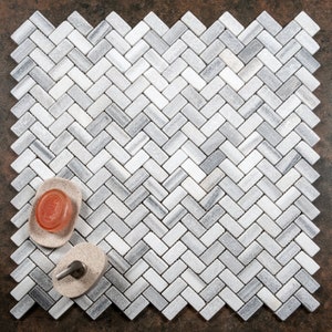 Può includere: Un mosaico di piastrelle di marmo bianco e grigio a spina di pesce. Due piccoli porta sapone rotondi beige sono sul mosaico. Un porta sapone contiene una saponetta rosa.