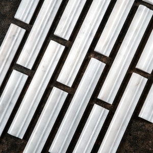May include: White marble tile trim pieces arranged in a diagonal pattern on a dark background. The tiles have a smooth, polished finish and a subtle raised edge.