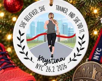 May include: A round white ceramic ornament featuring a runner crossing a finish line in a city. Text reads: SHE BELIEVED, SHE TRAINED, AND SHE DID IT. Krystina NYC 26.2 2026. The design includes black laurel branches and a gold hanging cord.