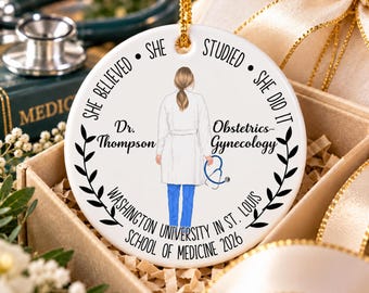 May include: A round white ceramic ornament featuring a central illustration of a doctor in a white coat and blue scrubs holding a stethoscope. Text reads: She believed she studied she did it, Dr. Thompson, Obstetrics-Gynecology, Washington University in St. Louis School of Medicine 2026.