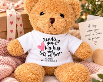 May include: A tan plush teddy bear wearing a white t-shirt with the text "sending you a big hug and lots of love" and "HUG ME BACK WHEN YOU ARE MISSING ME" printed in black, accompanied by a small pink heart graphic on the front of the shirt.