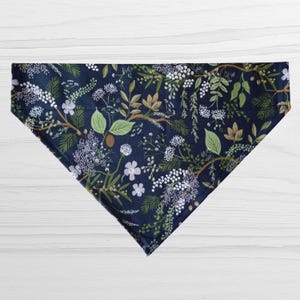 May include: A navy blue bandana with a botanical pattern. The design features white, green, and tan floral and foliage elements. The bandana is triangular in shape and made of fabric.