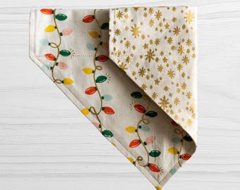 May include: A triangular pet bandana featuring a cream-colored fabric with a pattern of colorful Christmas light bulbs connected by thin brown lines, and a folded corner section displaying a gold metallic star print on a cream background.