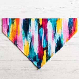 May include: A colorful dog bandana with a bright abstract pattern in shades of pink, yellow, blue, and red. The bandana is made of fabric and has a triangular shape.
