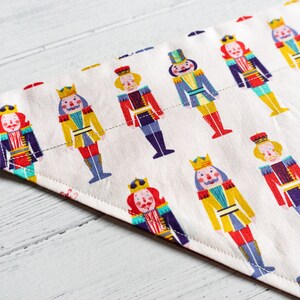 May include: A white fabric dog bandana with a repeating pattern of colorful nutcracker figures. The bandana is triangular in shape with a white background and a stitched edge.