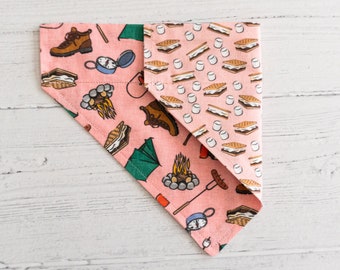 May include: Pink bandana with a camping theme. One side features a pattern of s'mores, while the other side has a pattern of camping gear, including a compass, boots, a tent, and a campfire.