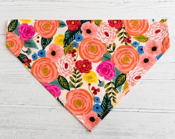 Puede incluir: Un pañuelo de algodón blanco con estampado floral con flores rosas, rojas, amarillas y azules. El pañuelo tiene forma de triángulo.