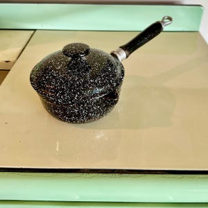 May include: A black and white speckled saucepan with a lid and a long, black handle. The small pot is sitting on a light green and cream-colored surface. The handle is attached with a silver metal piece.