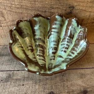 May include: A ceramic seashell-shaped dish with a glossy finish. The dish features a light green interior with brown accents along the edges and ridges. The dish is placed on a wooden surface.