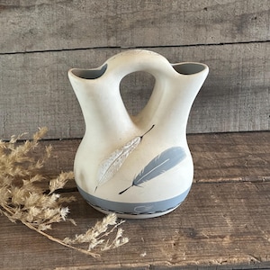 May include: Cream-colored ceramic vase with a unique double-spout design and a central handle. The vase features two feather designs in gray and white, with a gray band at the base. The vase is set against a rustic wooden background.