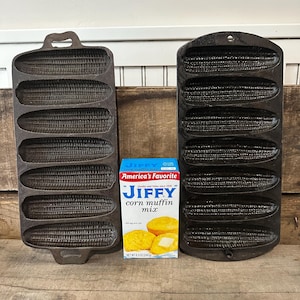 May include: Two cast iron corn-shaped muffin pans and a box of Jiffy corn muffin mix. The pans are black and have detailed corn cob impressions. The Jiffy box is blue, red, and white, with an image of baked muffins.