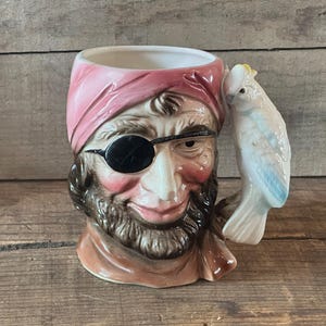May include: A ceramic character mug shaped like a pirate wearing a pink headscarf and a black eye patch. The mug features a brown beard and a sculpted white cockatoo with a yellow crest serving as the handle, set against a neutral background.