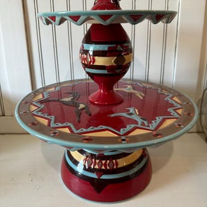 May include: A two-tiered ceramic cake stand with a Southwestern design. The stand features a red base and top with turquoise and brown accents. The top tier has a scalloped edge and a small decorative element on top.