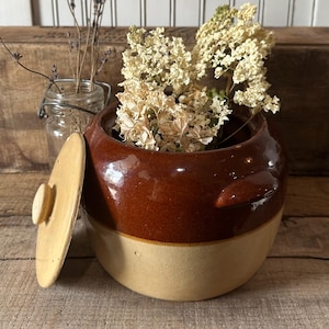 May include: A two-toned, brown and beige ceramic crock with a lid, filled with dried flowers. The crock has a glossy finish and two side handles. The lid is off to the side. A small glass jar with dried lavender is in the background.