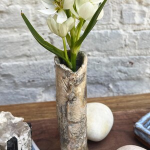 May include: A tall, cylindrical ceramic vase with a textured, mottled beige and grey finish holds a cluster of white star-shaped flowers with green stems and leaves. The vase has a rough, uneven rim and a weathered, earthy appearance.
