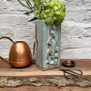 May include: A tall, rectangular, light blue ceramic vase with a textured, raised relief pattern, containing green flowers. Beside it sits a polished copper watering can, a small dark brown ceramic dish, and black metal floral shears.