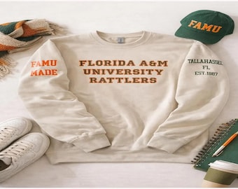 May include: Cream-colored sweatshirt with "FLORIDA A&M UNIVERSITY RATTLERS" in brown lettering. The left sleeve says "FAMU MADE" in orange, and the right sleeve says "TALLAHASSEE FL EST. 1887". A green hat with "FAMU" in gold, white sneakers, and a scarf are also visible.