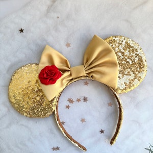 May include: Gold sequined Minnie Mouse ears with a large gold satin bow and a red rose accent.