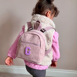 May include: A small, pale pink corduroy backpack with a purple letter "G" patch. The backpack features a zipped front pocket and adjustable straps. The child is wearing a pink jacket and a cream-coloured vest.