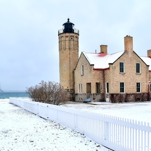 May include: A white picket fence in front of a brick lighthouse with a red roof. The lighthouse is on a snowy hill overlooking a body of water with a bridge in the distance.